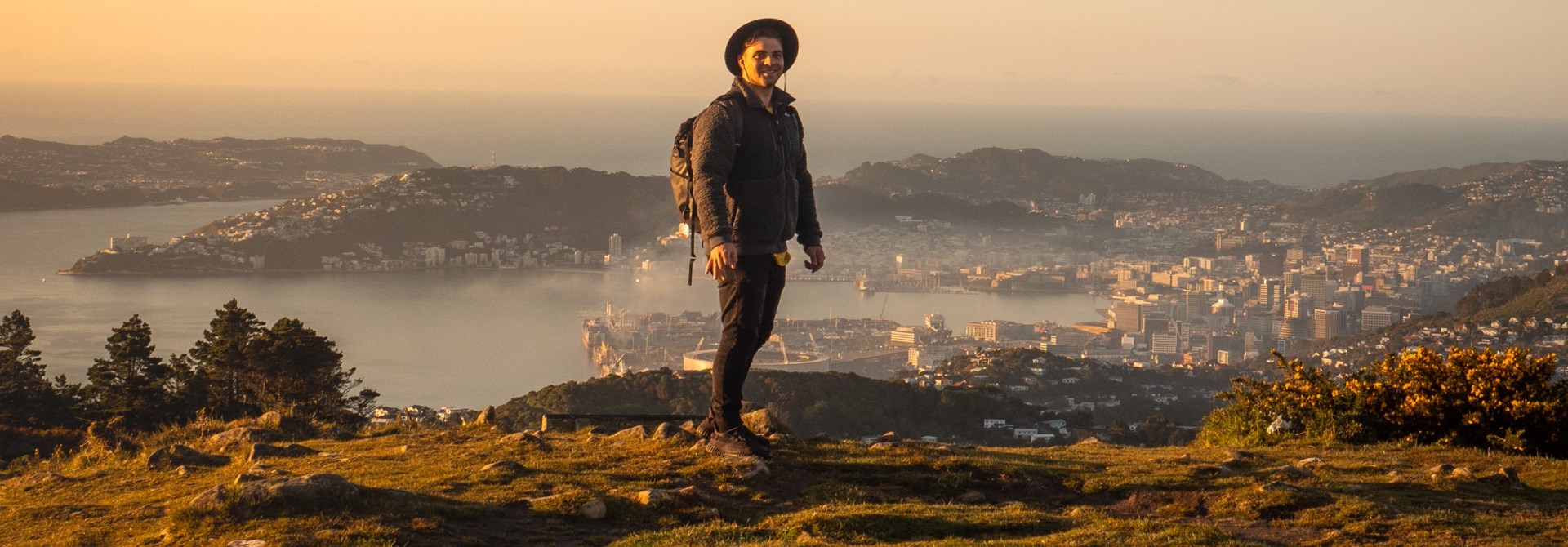Photographer Johnny Hendrikus standing on a hill as the sun goes down with mist around Wellington hills and harbour in background.